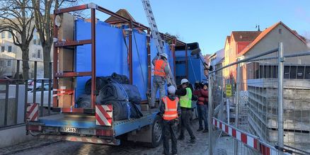 Blick auf einem LKW mit in blauer Folie eingepackten Aquarienscheiben auf der Ladefläche. Anbei stehen Arbeitskräfte in Baustellenbekleidung mit einer Leiter.
