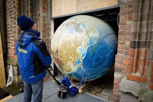 Ein Mann zieht einen großen Globus auf einem Transportmittel durch eine Türöffnung eines Backsteingebäudes. 