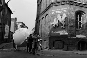 Schwarz-weiß-Bild auf dem Männer einen verhüllten Globus durch eine Straße tragen. An einer Hauswand hängt ein Plakat für das Meereskundliche Museum und Aquarium.  